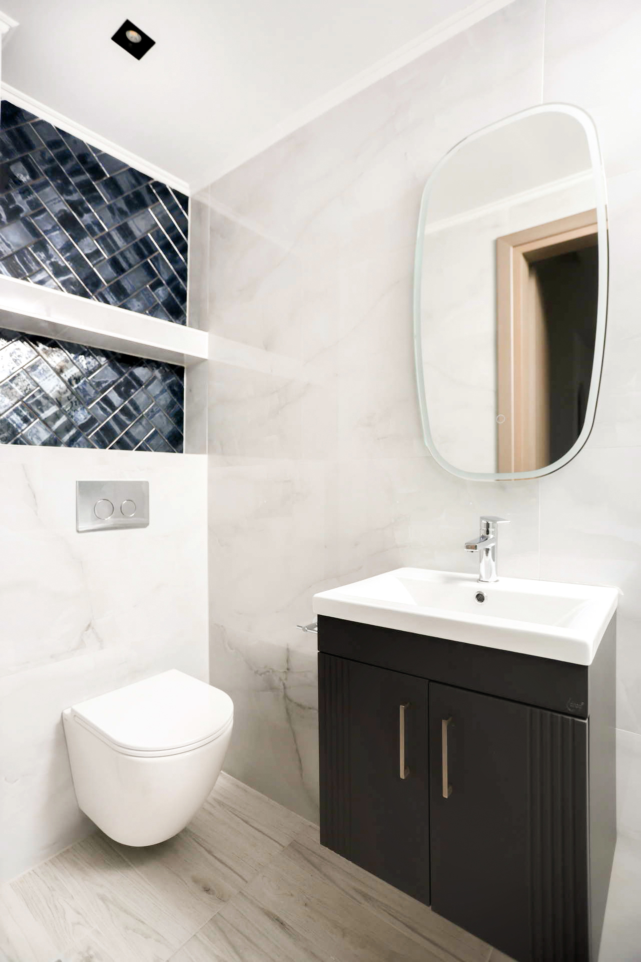 Modern bathroom with wall-mounted toilet, dark vanity, oval mirror, white marble walls and blue mosaic accent.