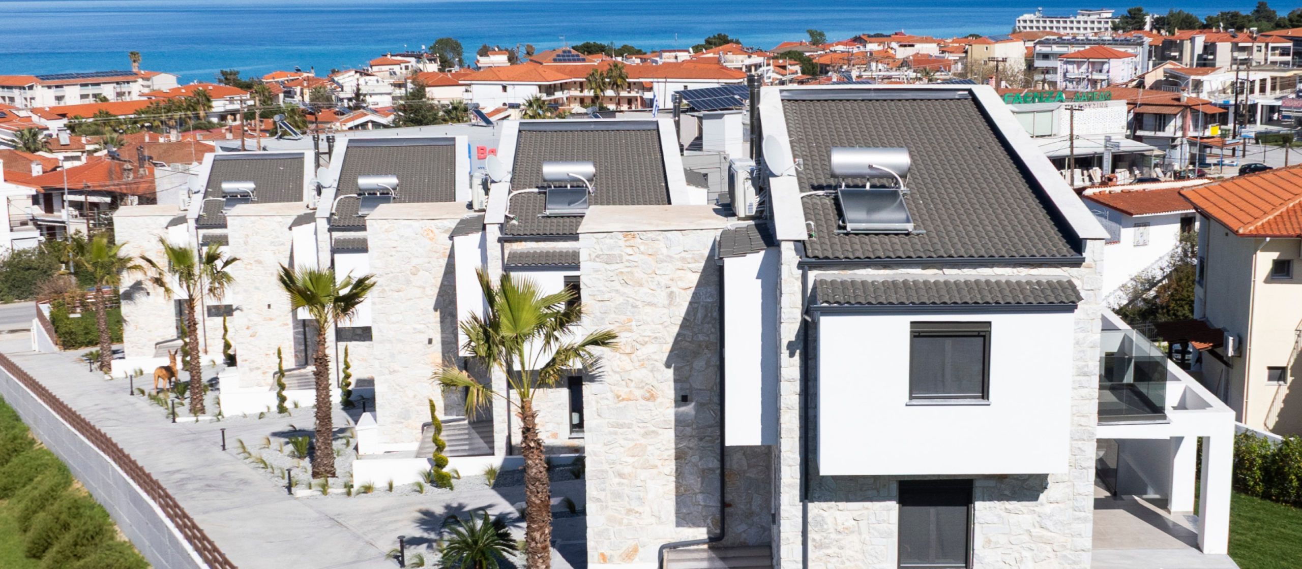 Coastal town with modern white stone villas, red-tiled roofs, palm trees, and sea view.
