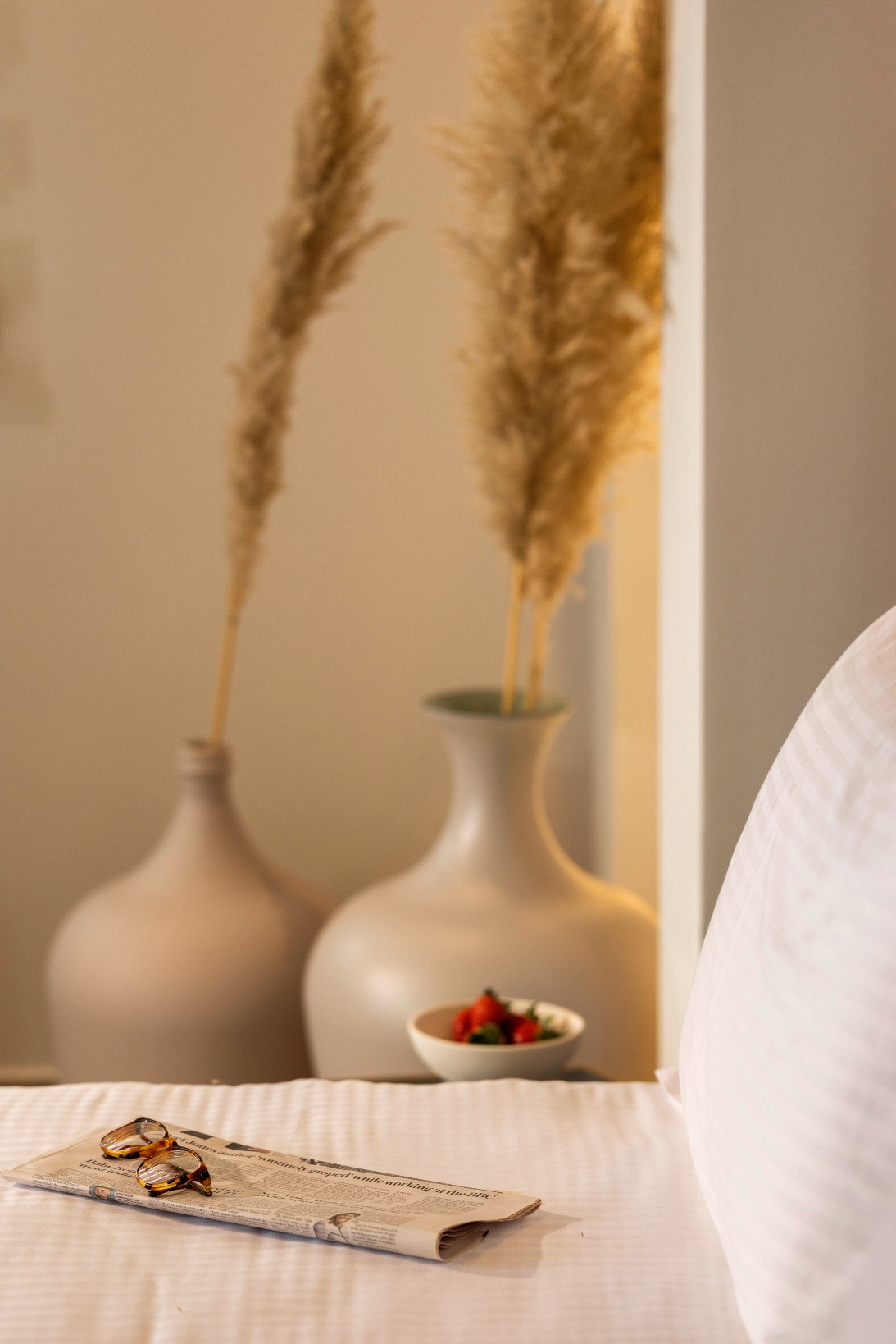 White bed with newspaper and glasses, small bowl of strawberries, and tall dried pampas grass in white vases.