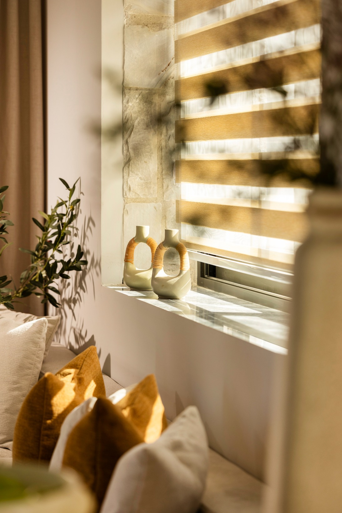 Sunlit living room window with striped blinds, two ceramic vases with woven orange handles, plant, and mustard cushions.