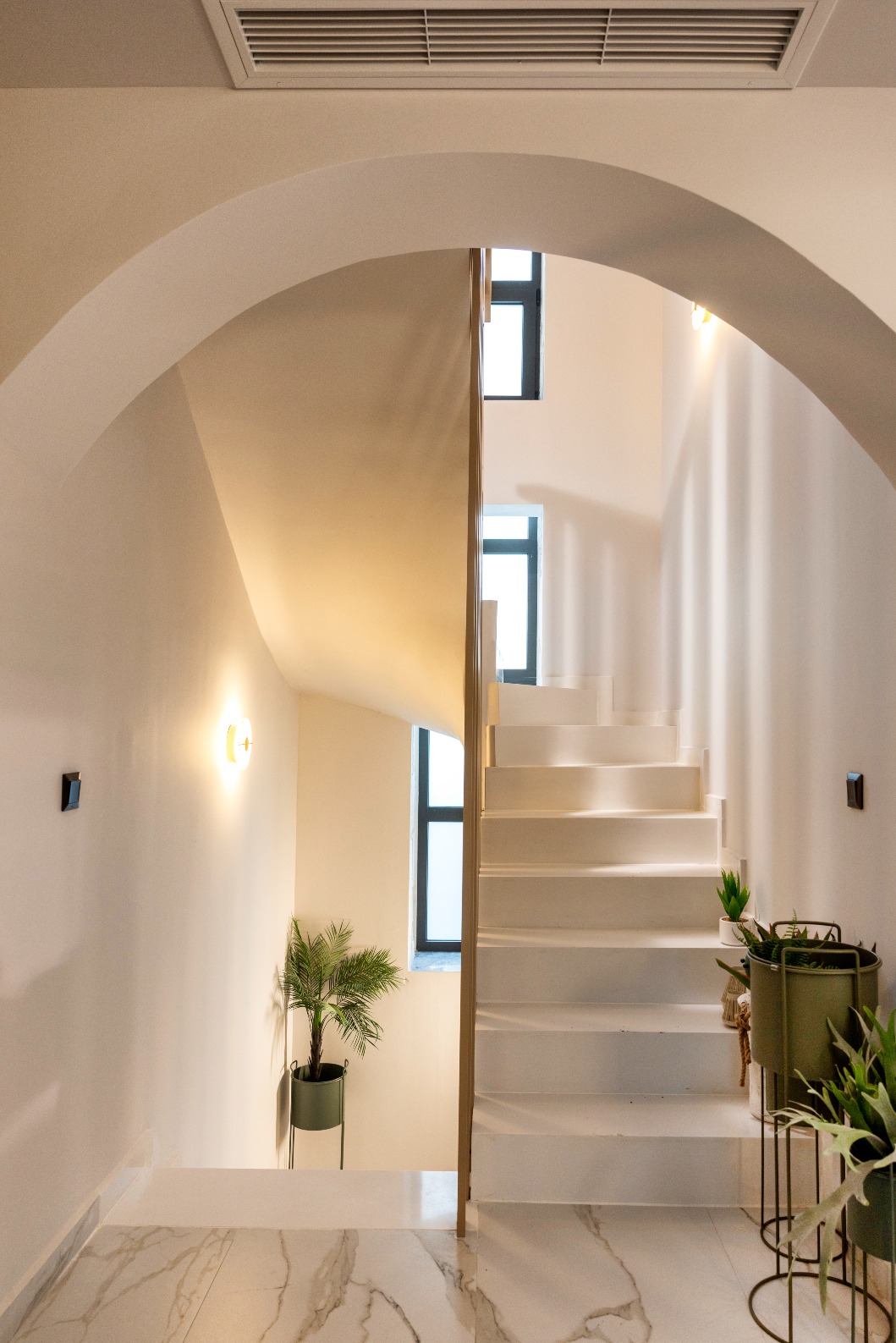 Modern white staircase with curved archway, marble floor, potted plants, and warm wall lights in a bright home interior.