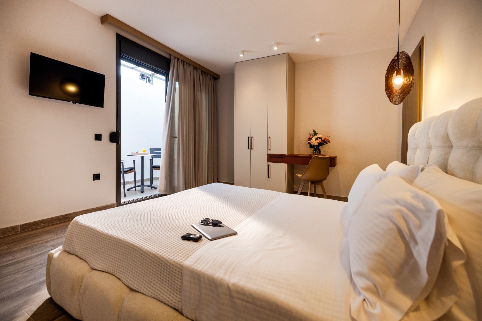 Modern hotel room with king bed, wall-mounted TV, balcony access, wardrobe, desk and warm pendant light.