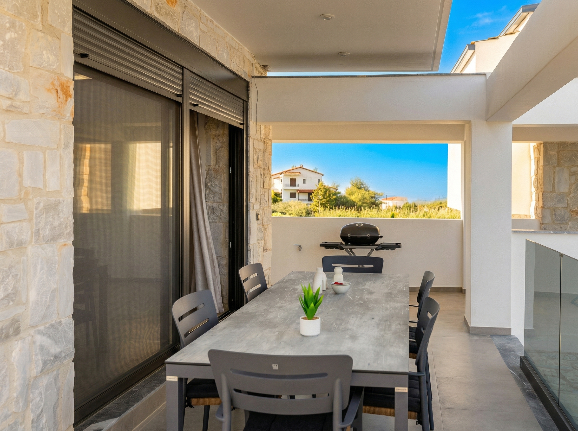 Outdoor terrace with stone walls, a long gray dining table, six chairs, and a grill in the background.