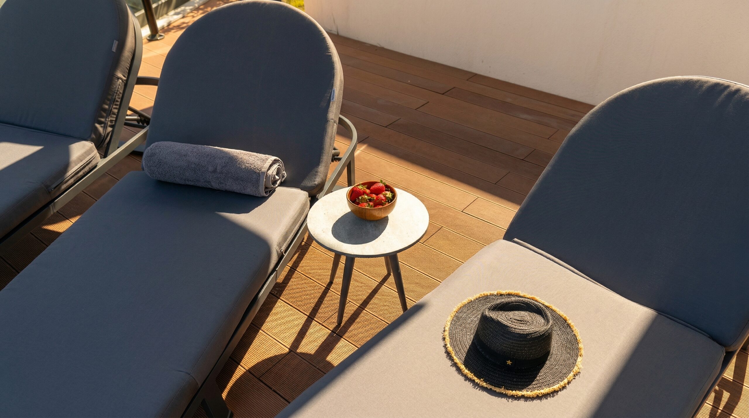 Outdoor wooden deck with blue lounge chairs, towel, marble table, bowl of strawberries, and a straw hat.