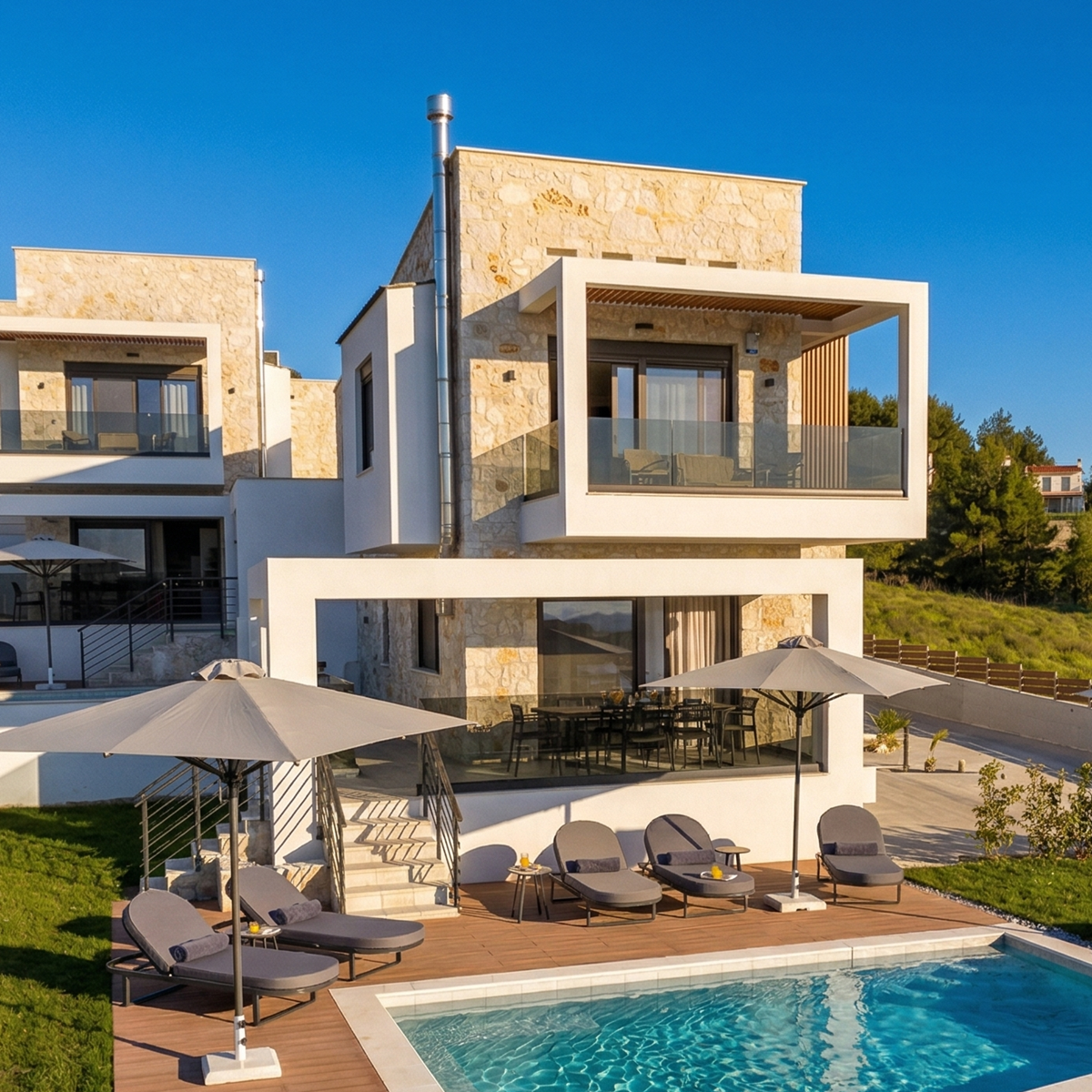 Modern two-storey villa in Greece Halkidiki with stone and white facades, large glass windows, and balconies. A patio with lounge chairs and umbrellas surrounds a rectangular swimming pool, set against a sunny, clear blue sky.