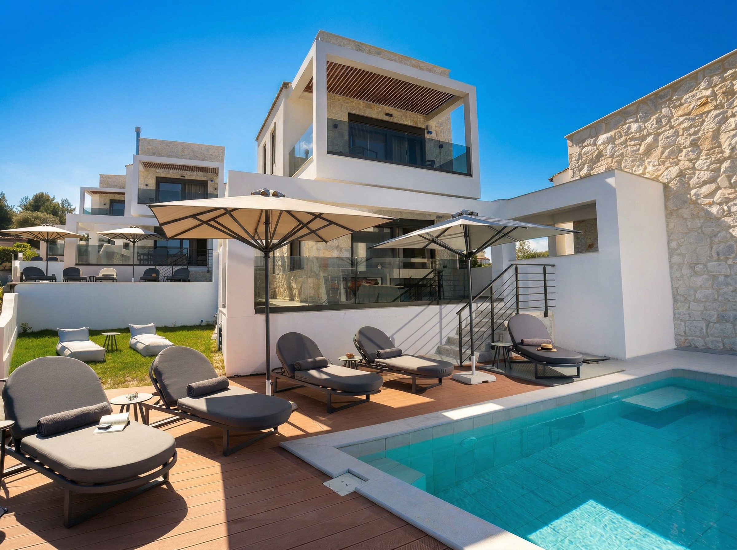 Modern villa with a private pool, sun loungers under umbrellas, glass balconies, white stone walls.