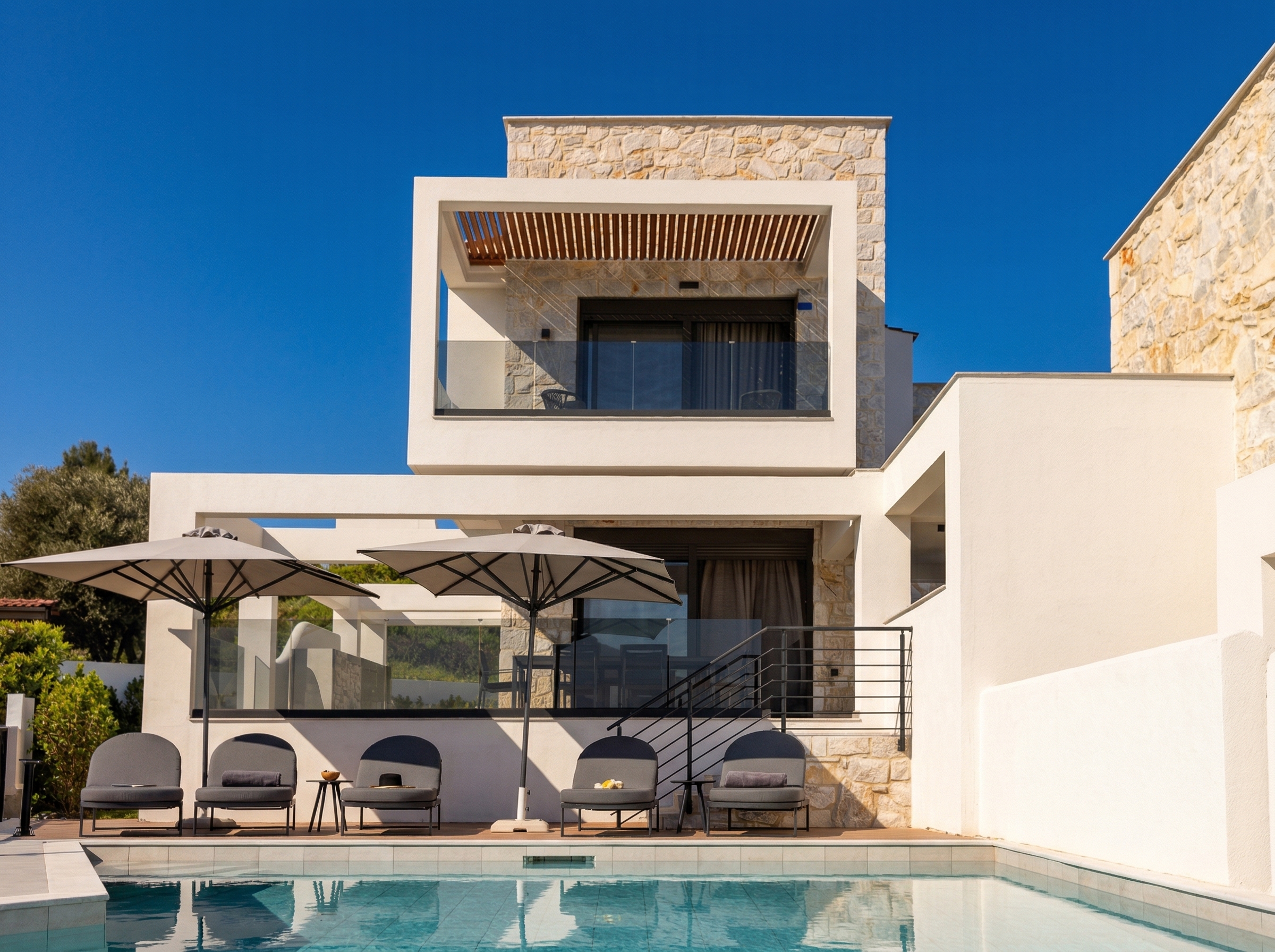 Modern luxury villa with stone facade, glass balcony, and poolside loungers under umbrellas.