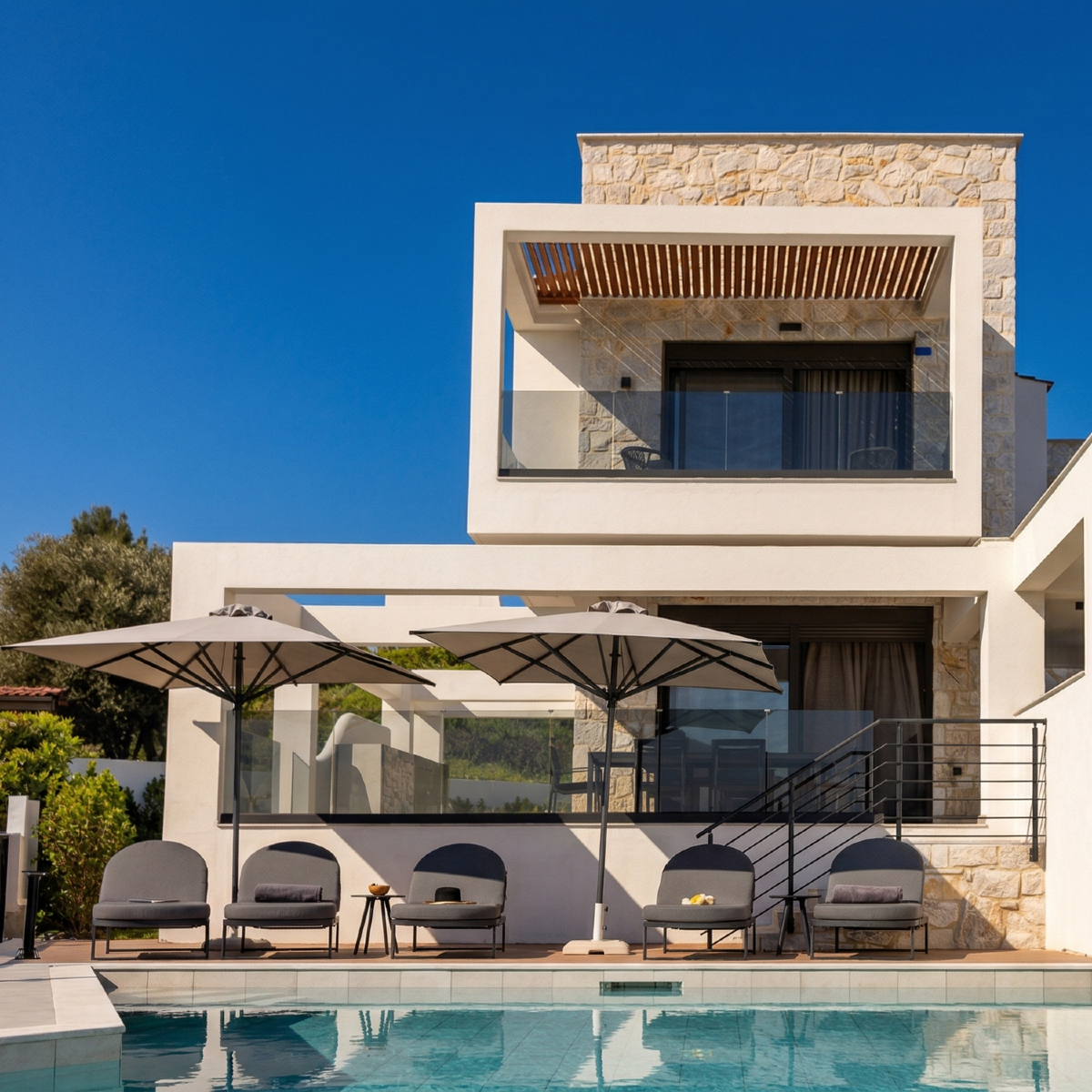 Modern villa in Greece Halkidiki with stone exterior, glass balcony, and poolside lounge under umbrellas.